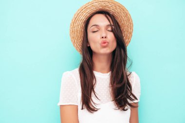 Moda kıyafetleri içinde gülümseyen güzel bir kadın. Stüdyoda mavi duvarın yanında poz veren kaygısız seksi kadın. Pozitif esmer model eğleniyor. Neşeli ve mutlu. Şapkalı. Ördek suratı yapar