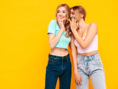 İki güzel, gülümseyen esmer hippi kadın moda yaz kıyafetleri içinde. Stüdyoda sarı duvarın yanında poz veren kaygısız seksi kadınlar. Pozitif model arkadaşına sırrını, dedikodusunu söyler.