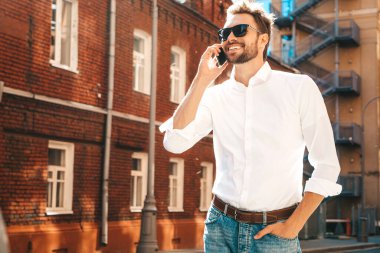 Yakışıklı, güler yüzlü, stil sahibi, lamberseksüel model. Beyaz gömlekli, modern bir adam. Sokakta güneş gözlükleriyle poz veren modacı erkek. Gün batımında dışarıda. Akıllı telefon kullanarak. Konuşarak.