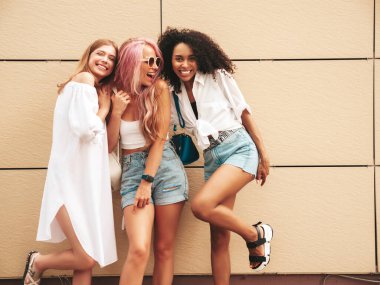 Son moda yaz kıyafetleri içinde gülümseyen üç genç bayan. Sokakta poz veren kaygısız, çok ırklı kadınlar. Güneş gözlükleriyle eğlenen pozitif modeller. Neşeli ve mutlu
