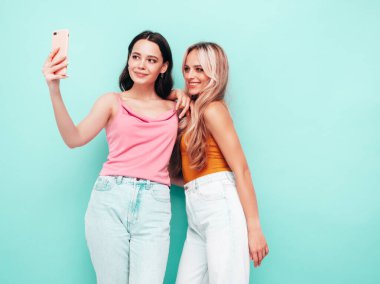 Moda kıyafetleri içinde gülümseyen iki güzel esmer hippi kadının portresi. Mavi duvarın yanında poz veren kaygısız seksi kadınlar. Pozitif modeller eğleniyor. Neşeli ve mutlu. selfie çekmek 