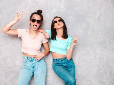 Moda kıyafetleri içinde gülümseyen iki güzel sarışın hippi kadın. Stüdyoda gri duvarın yanında poz veren kaygısız seksi kadınlar. Pozitif modeller eğleniyor. Neşeli ve mutlu. Güneş gözlükleriyle