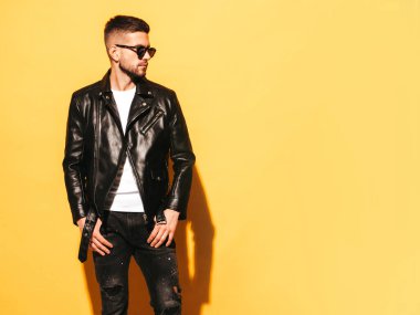 Yakışıklı, kendinden emin bir modelin portresi. Motosikletli deri ceket ve siyah kot giyen seksi şık bir adam. Moda hippi erkek. Stüdyoda güneş gözlüklü sarı duvarın yanında poz veriyor.