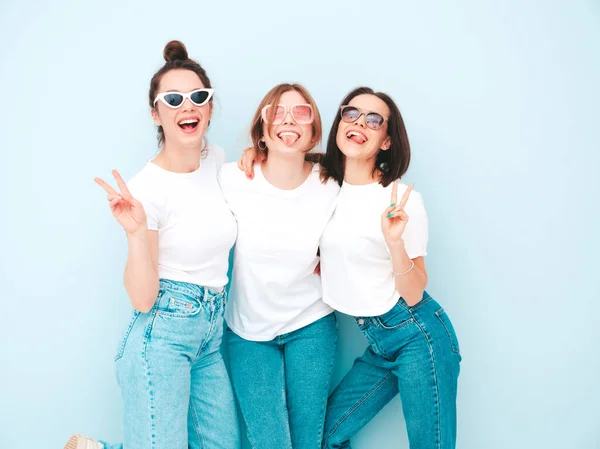三个年轻美丽的嬉皮士女 穿着时髦的夏天白色T恤和牛仔裤衣服 在演播室里 性感无忧无虑的女人在浅蓝色的墙壁附近摆姿势 快乐和积极的模型有乐趣 — 图库照片