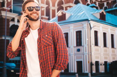 Yakışıklı, güler yüzlü, stil sahibi, lamberseksüel model. Kırmızı tişört giymiş modern bir adam. Sokakta güneş gözlükleriyle poz veren modacı erkek. Gün batımında dışarıda. Akıllı telefon kullanarak. Konuşarak.