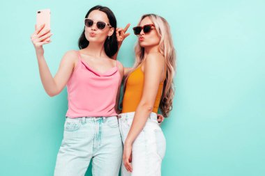 Moda kıyafetleri içinde gülümseyen iki güzel esmer hippi kadının portresi. Mavi duvarın yanında poz veren kaygısız seksi kadınlar. Pozitif modeller eğleniyor. Neşeli ve mutlu. selfie çekmek 