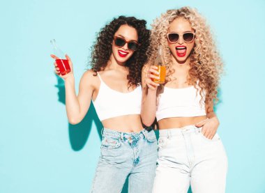 Yaz kıyafetleri içinde gülümseyen iki güzel hippi kadın. Saç stili kıvırcık saçlı moda modeller stüdyoda eğleniyor. Mavide izole edilmiş seksi bir kadın. Limonata fro cam şişesi içiyor. Güneş gözlükleriyle
