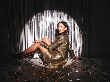 Güzelim, güler yüzlü, esmer kadın, son moda, altın ceketli. Stüdyoda gümüş renkli parlak bir duvarın yanında poz veren kaygısız seksi bir kadın. Parlak makyajlı, modaya uygun bir model. Yerde otururken.