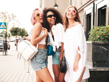 Son moda yaz kıyafetleri içinde gülümseyen üç genç bayan. Sokakta poz veren kaygısız, çok ırklı kadınlar. Güneş gözlükleriyle eğlenen pozitif modeller. Neşeli ve mutlu