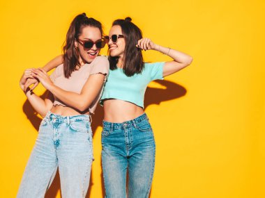 Moda kıyafetleri içinde gülümseyen iki güzel sarışın hippi kadın. Stüdyoda sarı duvarın yanında poz veren kaygısız seksi kadınlar. Pozitif modeller eğleniyor. Neşeli ve mutlu. Güneş gözlükleriyle