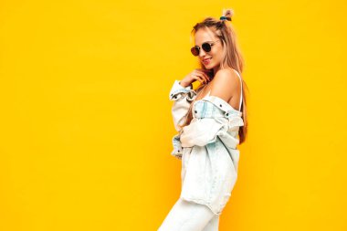 Moda kıyafetleri içinde gülümseyen sarışın güzel bir kadının portresi. Stüdyoda sarı duvarın yanında poz veren kaygısız seksi kadın. Pozitif model içeride eğleniyor. Neşeli ve mutlu güneş gözlükleriyle
