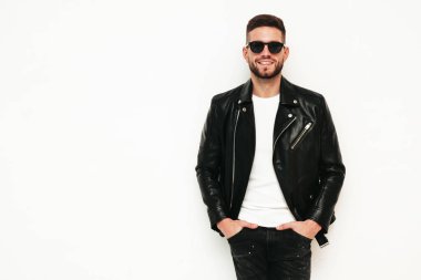 Yakışıklı gülümseyen modelin portresi. Motosikletli deri ceket ve siyah kot giyen seksi şık bir adam. Moda meraklısı erkek, stüdyoda beyazdan soyutlanmış. Güneş gözlükleriyle