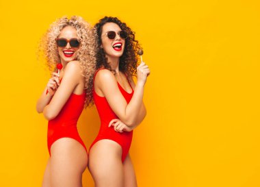 Kırmızı mayo giymiş iki güzel gülümseyen hippi kadın. Stüdyoda kıvırcık saçlı moda modeller. Güneş gözlüklü, sarı duvarın yanında poz veren seksi bir kadın. Lolipop tutuyor. Beyazda izole edilmiş