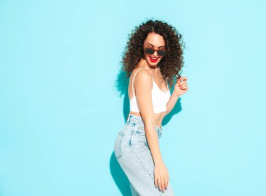 Yeni moda hippi kıyafetleri içinde gülümseyen güzel bir kadın. Stüdyoda mavi duvarın yanında poz veren kaygısız bir kadın. Kıvırcık saçlı esmer bir model. Neşeli ve mutlu. Güneş gözlükleriyle