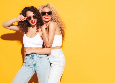 Yaz kıyafetleri içinde gülümseyen iki güzel hippi kadın. Saç stili kıvırcık saçlı moda modeller stüdyoda eğleniyor. Sarıda izole edilmiş seksi bir kadın. Mutlu ve neşeli. Güneş gözlükleriyle