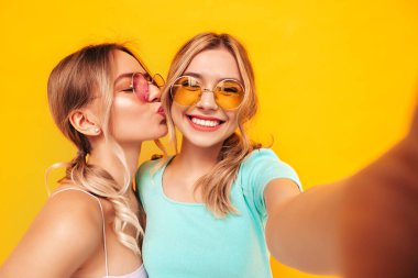 Moda kıyafetleri içinde gülümseyen iki güzel sarışın hippi kadın. Stüdyoda sarı duvarın yanında poz veren kaygısız seksi kadınlar. Pozitif modeller eğleniyor. Güneş gözlüklü. Pov 'un fotoğrafını çekiyorum.