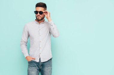 Yakışıklı, kendinden emin bir modelin portresi. Gömlek ve kot giyen seksi şık bir adam. Moda hippi erkek stüdyoda mavi duvarın yanında poz veriyor. Neşeli ve mutlu. Güneş gözlükleriyle