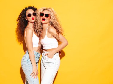 Yaz kıyafetleri içinde gülümseyen iki güzel hippi kadın. Saç stili kıvırcık saçlı moda modeller stüdyoda eğleniyor. Sarıda izole edilmiş seksi bir kadın. Mutlu ve neşeli. Güneş gözlükleriyle