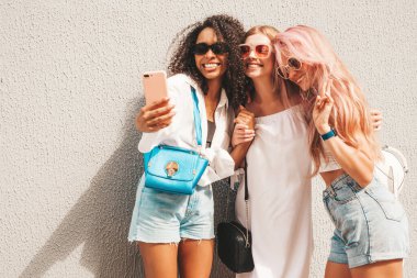 Son moda yaz kıyafetleri içinde üç güzel gülümseyen hippi kadın. Sokakta poz veren tasasız, çok ırklı kadınlar. Güneş gözlükleriyle eğlenen pozitif modeller. Selfie çekiyoruz.