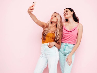 İki güzel, gülümseyen esmer hippi kadın moda yaz kıyafetleri içinde. Pembe duvarın yanında poz veren kaygısız seksi kadınlar. Pozitif modeller eğleniyor. Neşeli ve mutlu. Selfie çekiyoruz. 
