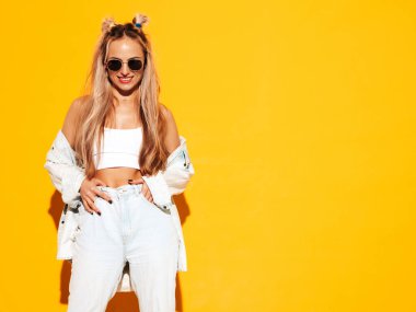 Moda kıyafetleri içinde gülümseyen sarışın güzel bir kadının portresi. Stüdyoda sarı duvarın yanında poz veren kaygısız seksi kadın. Pozitif model içeride eğleniyor. Neşeli ve mutlu güneş gözlükleriyle