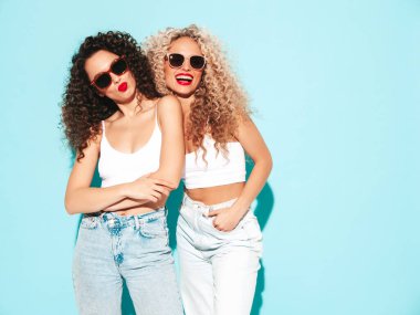 Yaz kıyafetleri içinde gülümseyen iki güzel hippi kadın. Saç stili kıvırcık saçlı moda modeller stüdyoda eğleniyor. Mavide izole edilmiş seksi bir kadın. Mutlu ve neşeli. Güneş gözlükleriyle