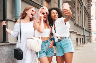 Son moda yaz kıyafetleri içinde üç güzel gülümseyen hippi kadın. Sokakta poz veren tasasız, çok ırklı kadınlar. Güneş gözlükleriyle eğlenen pozitif modeller. Selfie çekiyoruz.