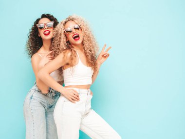 Yaz kıyafetleri içinde gülümseyen iki güzel hippi kadın. Saç stili kıvırcık saçlı moda modeller stüdyoda eğleniyor. Mavide izole edilmiş seksi bir kadın. Mutlu ve neşeli. Güneş gözlükleriyle