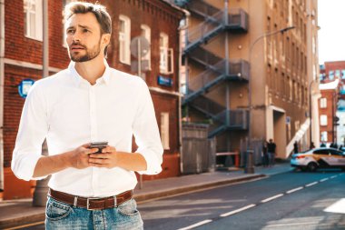 Yakışıklı, güler yüzlü, stil sahibi, lamberseksüel model. Beyaz gömlekli, modern bir adam. Moda erkeği sokakta poz veriyor. Gün batımında dışarıda. Akıllı telefon uygulamaları kullanarak. Cep telefonu tutuluyor