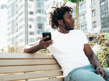 Yakışıklı hippi mankeni. Beyaz yaz tişörtü giymiş, traşsız Afrikalı bir adam. Sokakta bankta oturan rastalı saçlı moda bir erkek.