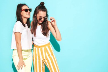 Moda kıyafetleri içinde gülümseyen iki güzel sarışın hippi kadın. Stüdyoda mavi duvarın yanında poz veren kaygısız seksi kadınlar. Pozitif modeller eğleniyor. Neşeli ve mutlu