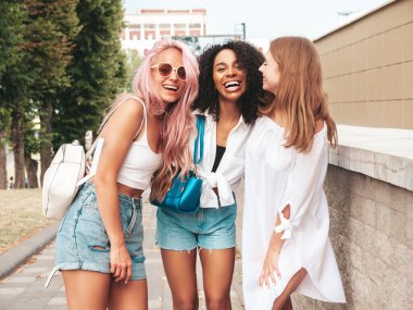 Son moda yaz kıyafetleri içinde gülümseyen üç genç bayan. Sokakta poz veren kaygısız, çok ırklı kadınlar. Güneş gözlükleriyle eğlenen pozitif modeller. Neşeli ve mutlu