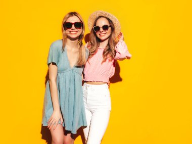 İki güzel, gülümseyen esmer hippi kadın moda yaz elbiseleri içinde. Sarı duvarın yanında poz veren kaygısız seksi kadınlar. Pozitif modeller eğleniyor. Neşeli ve mutlu. Şapka ve güneş gözlükleriyle
