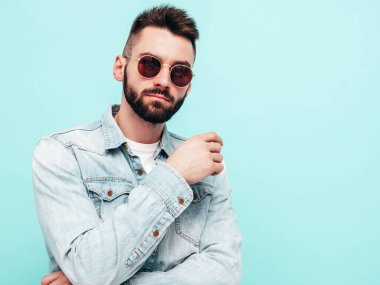 Yakışıklı, kendine güvenen, stil sahibi, Lambersexual modelin portresi. Ceket ve kot giyen adam. Moda erkeği, stüdyoda güneş gözlüklü mavi duvarın yanında poz veriyor.