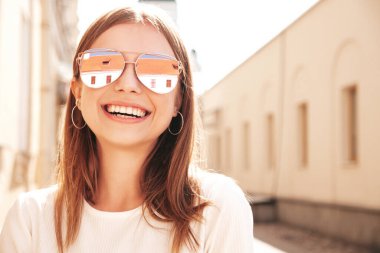 Moda kıyafetleri içinde gülümseyen genç ve güzel bir hippi kadın. Günbatımında sokakta poz veren kaygısız seksi kadın. Açık havada pozitif model. Neşeli ve mutlu güneş gözlükleriyle