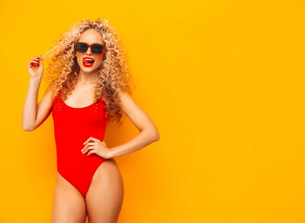 スタジオで黄色の壁の近くにポーズ若い美しい笑顔の女性 赤い水着水着でセクシーなモデルは それを水着 カールヘアスタイルと正の女性 サングラスで — ストック写真