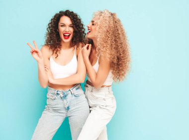 Yaz kıyafetleri içinde gülümseyen iki güzel hippi kadın. Saç stili kıvırcık saçlı moda modeller stüdyoda eğleniyor. Mavide izole edilmiş seksi bir kadın. Mutlu ve neşeli. Güneş gözlükleriyle