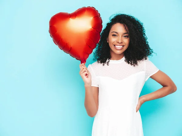 Beautiful black woman with afro curls hairstyle. Smiling model dressed in white summer dress. Sexy carefree female posing near blue wall in studio. Tanned and cheerful. Holding heart air balloon