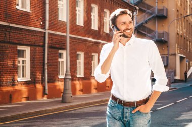 Yakışıklı, güler yüzlü, stil sahibi, lamberseksüel model. Beyaz gömlekli, modern bir adam. Sokakta güneş gözlükleriyle poz veren modacı erkek. Gün batımında dışarıda. Akıllı telefon kullanarak. Konuşarak.