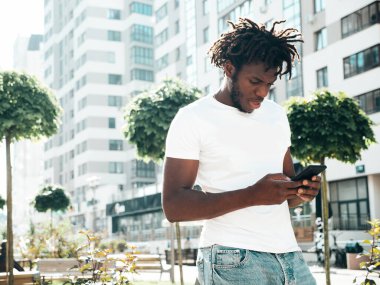 Yakışıklı hippi mankeni. Beyaz yaz tişörtü giymiş traşsız Afrikalı bir adam. Rasta saçlı, moda bir erkek. Sokakta poz vermek. Akıllı telefon ekranına bakmak, cep telefonu uygulamaları kullanmak.