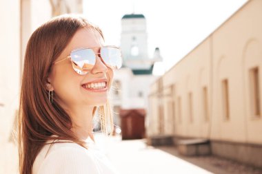 Moda kıyafetleri içinde gülümseyen genç ve güzel bir hippi kadın. Günbatımında sokakta poz veren kaygısız seksi kadın. Açık havada pozitif model. Neşeli ve mutlu güneş gözlükleriyle