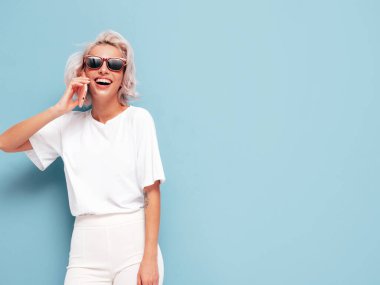 Moda kıyafetleri içinde gülümseyen güzel bir kadın. Stüdyoda mavi duvarın yanında poz veren kaygısız seksi kadın. Pozitif sarışın model cep telefonuyla konuşuyor. Akıllı telefonunu kullanarak ve konuşarak.