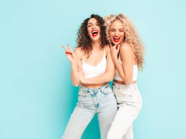 Yaz kıyafetleri içinde gülümseyen iki güzel hippi kadın. Saç stili kıvırcık saçlı moda modeller stüdyoda eğleniyor. Mavide izole edilmiş seksi bir kadın. Mutlu ve neşeli. Güneş gözlükleriyle