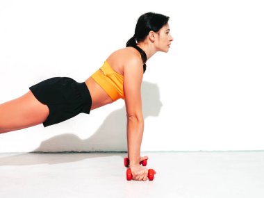 Siyah spor elbiseli kendine güvenen bir kadın. Mükemmel vücudu olan genç ve seksi bir manken. Stüdyoda beyaz duvarda izole edilmiş bir kadın var. Eğitimden önce esneme hareketleri. Dambıllara basarak.