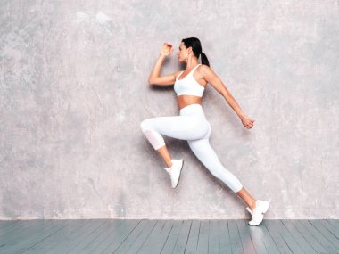 Seksi spor elbiseli spor kadını. Mükemmel vücutlu genç ve güzel bir model. Güneşli yaz gününde gri duvarın yanındaki stüdyoda poz veren bir kadın. Zıplıyor ve koşuyor. Koşuyorum. 