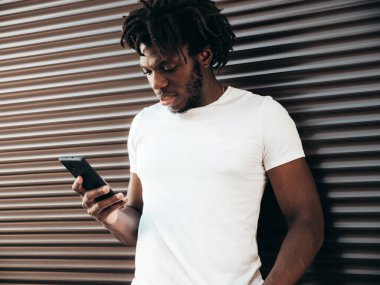Yakışıklı hippi mankeni. Beyaz yaz tişörtü giymiş traşsız Afrikalı bir adam. Rasta saçlı, moda bir erkek. Sokakta poz vermek. Akıllı telefon ekranına bakmak, cep telefonu uygulamaları kullanmak.