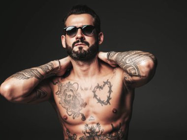 Yakışıklı, kendine güvenen, stil sahibi hipster Lambersex modelinin portresi. Seksi, modern adam. Dövmeli çıplak gövde. Stüdyoda güneş gözlüklü, siyah arka planda poz veren moda erkek.