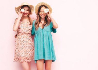 İki güzel, gülümseyen esmer hippi kadın moda yaz elbiseleri içinde. Pembe duvarın yanında poz veren kaygısız seksi kadınlar. Çiçek tutan pozitif modeller. Neşeli ve mutlu. Şapkalı. 