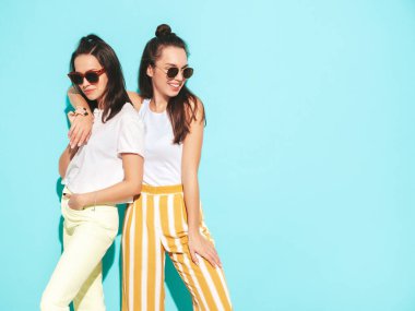 Moda kıyafetleri içinde gülümseyen iki güzel sarışın hippi kadın. Stüdyoda mavi duvarın yanında poz veren kaygısız seksi kadınlar. Pozitif modeller eğleniyor. Neşeli ve mutlu