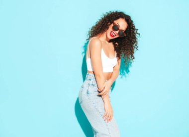 Yeni moda hippi kıyafetleri içinde gülümseyen güzel bir kadın. Stüdyoda mavi duvarın yanında poz veren kaygısız bir kadın. Kıvırcık saçlı esmer bir model. Neşeli ve mutlu. Güneş gözlükleriyle